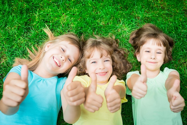 Glückliche Kinder, die im Frühlingspark auf grünem Gras liegen Lachende Kinder, die Daumen nach oben zeigen