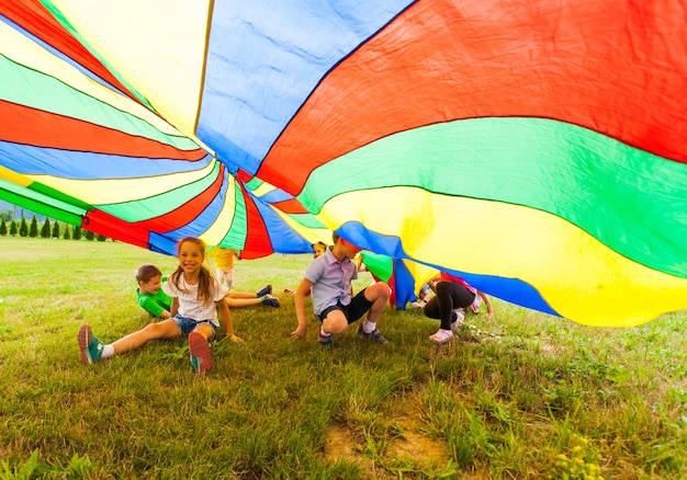 Glückliche Kinder, die sich draußen unter einem riesigen Regenbogenfallschirm verstecken