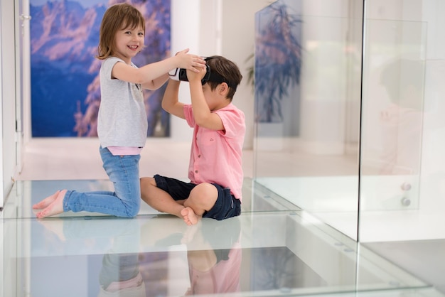 Glückliche Kinder, die Virtual-Reality-Headsets verwenden und mit dem Finger nach oben zeigen, während sie auf dem Boden in einem modernen, hellen Wohninterieur sitzen