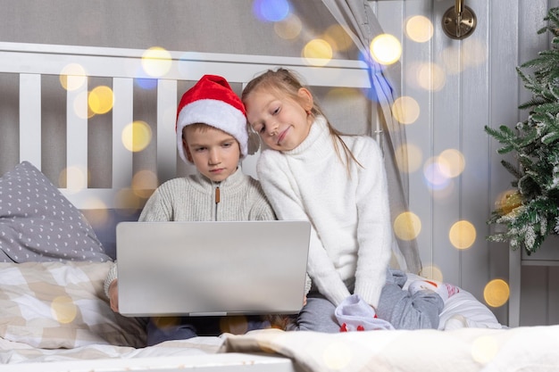 Glückliche Kinder in roten Weihnachtsmützen mit einem Laptop an Heiligabend zu Hause Ein Junge und ein Mädchen gratulieren Verwandten online