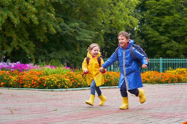 Glückliche Kinder lachen in Eile und laufen in Regenmänteln mit einer Aktentasche hinter einem Rucksack zur Schule.