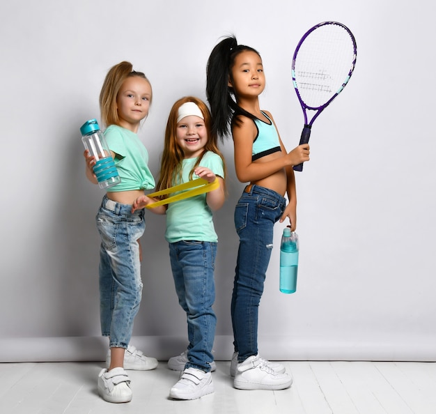 Glückliche Kinder mit Sportgerät-Studioaufnahmeporträt lokalisiert auf weißem Hintergrund. Kleine multiethnische Mädchen in Jeans, sportliche T-Shirts halten Wasserflasche, großen Tennisschläger, Fitnessgummi fitness