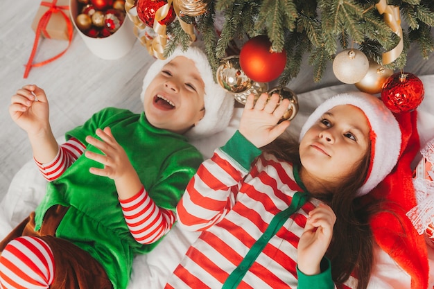 Glückliche Kinderjunge und -mädchen liegen auf dem Boden nahe einem festlich geschmückten Weihnachtsbaum. Weihnachten