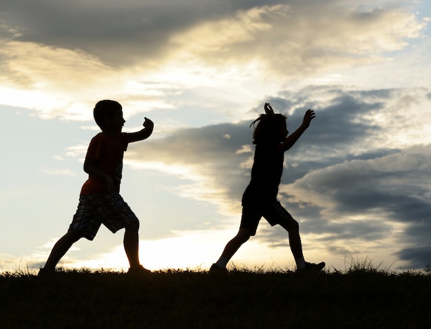 Glückliche Kinderschattenbilder, die Spaß auf Wiese bei dem Sonnenuntergang feiert Sommer haben