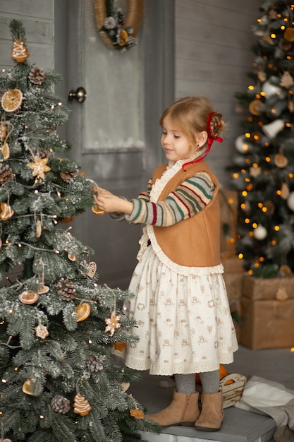 Glückliche kleine Blondine in Weihnachtskleidung, die den Weihnachtsbaum schmückt und lächelt