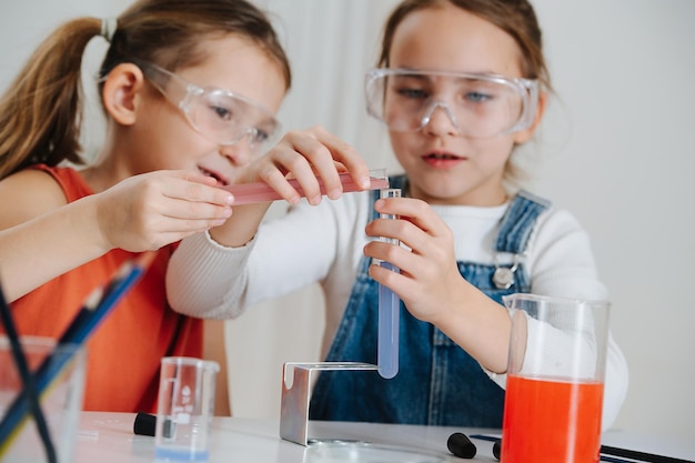 Glückliche kleine Mädchen, die ein Wissenschaftsprojekt machen, mischen zwei farbige Flüssigkeiten und gießen von einer Flasche in eine andere. Beide tragen eine Schutzbrille.