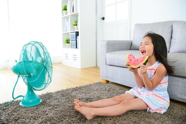 glückliche kleine mädchen sitzen auf dem boden des wohnzimmers, blasen einen elektrischen ventilator, kühlen wind und essen frische wassermelone, die sich im sommer wohl fühlen.