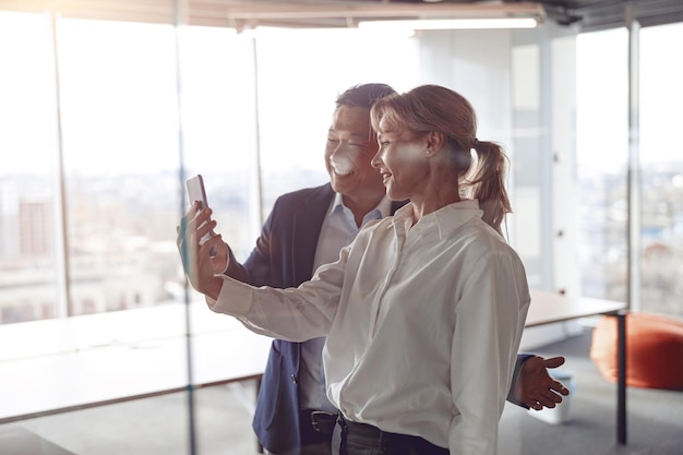 Glückliche Kollegen nutzen Smartphones für Videoanrufe, während sie in einem modernen Büro in der Nähe einer Glaswand stehen