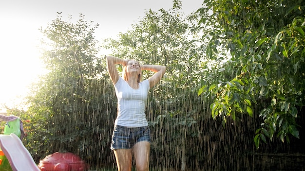 Glückliche lachende junge Frau, die unter Regen im Garten tanzt. Mädchen, das im Sommer im Freien spielt und Spaß hat