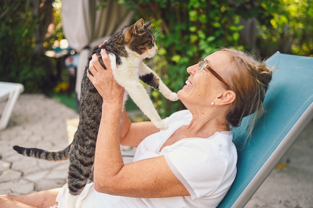 Glückliche lächelnde ältere ältere Frau in den Gläsern, die im Sommergarten im Freien entspannen, der inländische Tabbykatze umarmt. Pensioniertes altes Menschen- und Tierhaustierkonzept