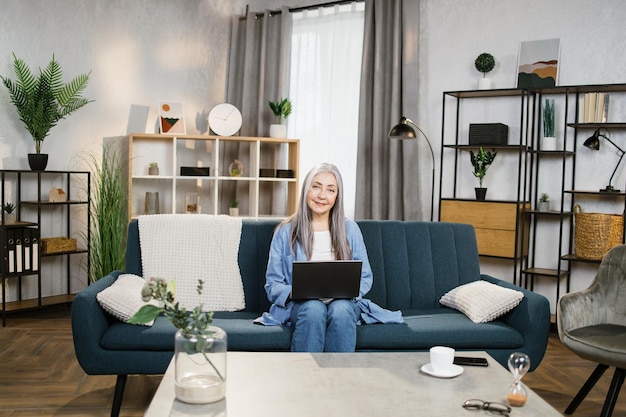 Glückliche lächelnde ältere Frau, die auf blauem Sofa sitzt und zu Hause mit Laptop studiert