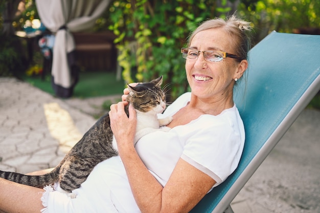 Glückliche lächelnde ältere Frau in den Gläsern, die im Sommergarten draußen entspannen, inländische Tabbykatze umarmend