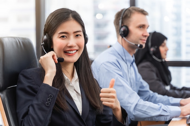 Glückliche lächelnde asiatische Frau des Betreibers ist Kundendienstmitarbeiter mit den Kopfhörern, die an Computer in einem Call-Center arbeiten und spricht mit Kunden, damit die Unterstützung das Problem mit den Haltungsdaumen oben löst