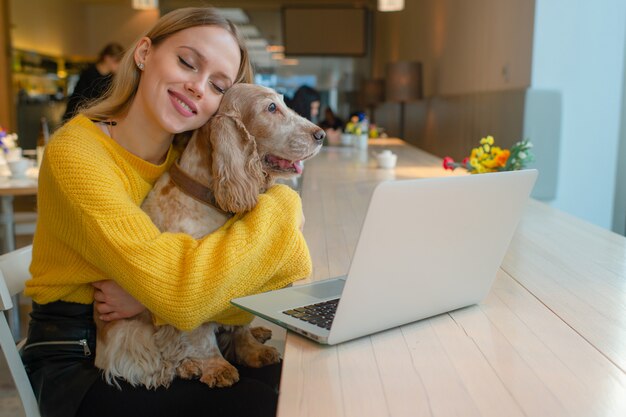 Glückliche lächelnde blonde Frau, die ihren schönen Cockerspanielwelpen vor Laptop auf dem Tisch im Kaffee umarmt.