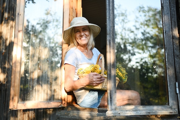Glückliche lächelnde emotionale ältere Frau, die durch offenes Fenster im alten hölzernen Dorfhaus im Strohhut mit Blumenkorb aufwirft