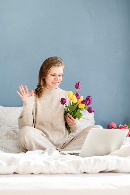 Glückliche lächelnde Frau, die auf dem Bett sitzt und Pyjamas trägt, mit Vergnügen Blumen genießt und am Laptop arbeitet