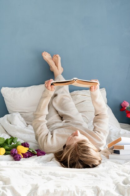 Glückliche lächelnde Frau, die auf dem Bett sitzt und Pyjamas trägt, mit Vergnügen Blumen genießt und ein Buch liest