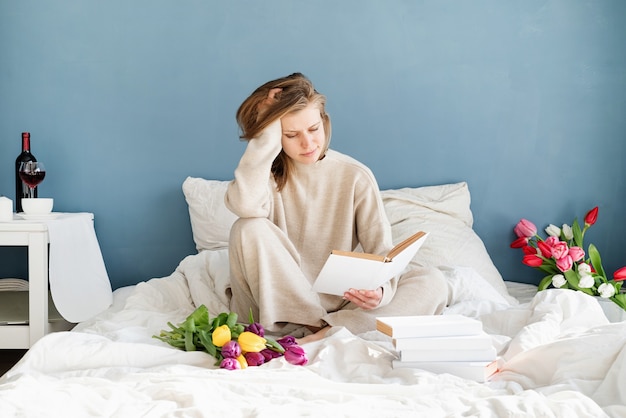Glückliche lächelnde Frau, die auf dem Bett sitzt und Pyjamas trägt, mit Vergnügen, Blumen zu genießen und ein Buch zu lesen