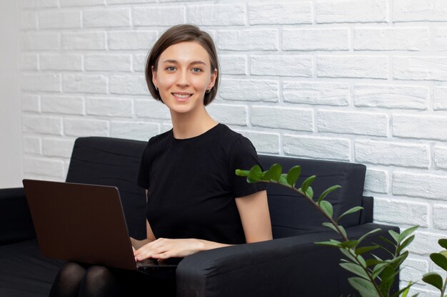 Glückliche lächelnde Frau, die auf Laptop-Computer vom Büro sitzt, das auf Sofa sitzt