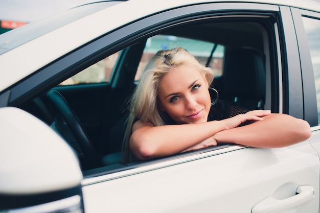 Glückliche lächelnde Frau, die sich über neuen teuren Kauf im Autohaus freut. Schöner Kunde, der auf Tür des weißen Faltverdeckautos sitzt.