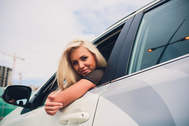 Glückliche lächelnde Frau, die sich über neuen teuren Kauf im Autohaus freut. Schöner Kunde, der auf Tür des weißen Faltverdeckautos sitzt.