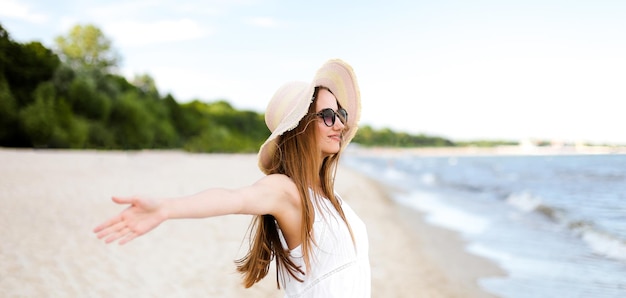 Glückliche lächelnde Frau in freier Glückseligkeit auf dem Ozeanstrand mit Hut, Sonnenbrille und offenen Händen. Porträt eines multikulturellen weiblichen Models in weißem Sommerkleid, das während einer Reise die Natur genießt