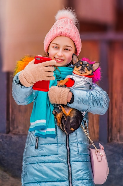 Glückliche lächelnde Frau macht am Wintertag Selbstporträt auf dem Smartphone. Jugendlichmädchen und Chihuahua.