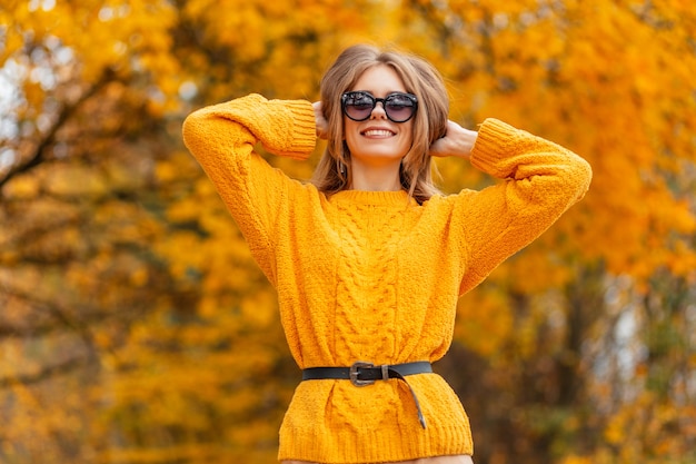 Glückliche lächelnde Frau mit modischer Sonnenbrille im gelben Strickpullover genießen Sie einen Herbsttag mit goldenen Blättern