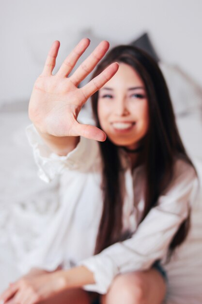 Glückliche lächelnde junge frau des schönen langen asiatischen mädchens des haares, die hand in richtung zur kamera sitzt auf bett zieht