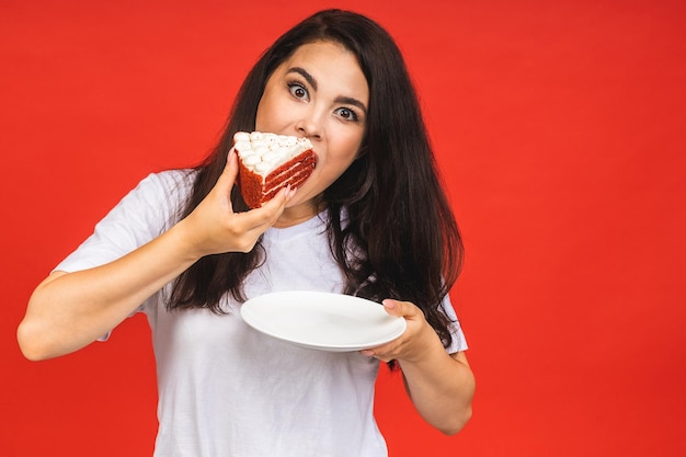 Glückliche lächelnde junge Frau, die den Kuchen isst, der über rotem Hintergrund lokalisiert wird Brunettedame, die einen Geburtstagskuchen hält