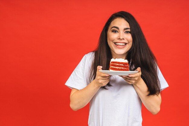 Glückliche lächelnde junge Frau, die den Kuchen isst, der über rotem Hintergrund lokalisiert wird Brunettedame, die einen Geburtstagskuchen hält