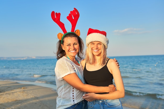 Glückliche lächelnde Mutter und jugendliche Tochter im Weihnachtsmannhut am Strand