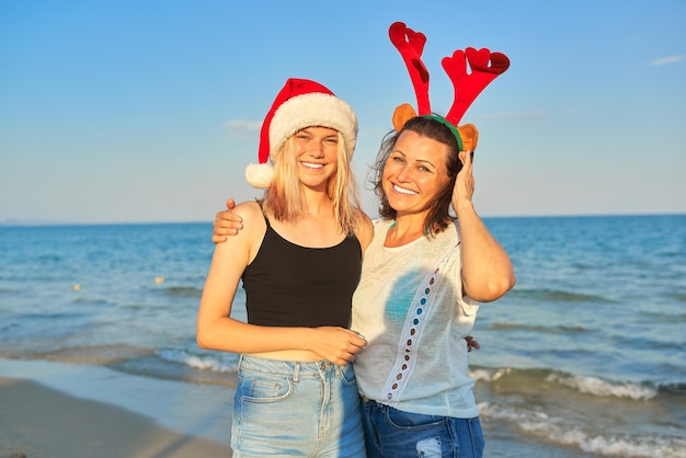 Glückliche lächelnde Mutter und jugendliche Tochter im Weihnachtsmannhut am Strand