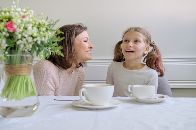 Glückliche lächelnde Mutter und kleine Tochter, die am Tisch der Tassen trinken