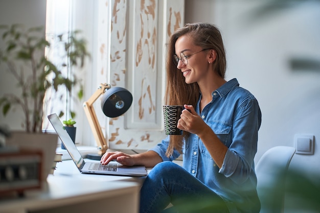 Glückliche lässige junge lächelnde Frau, die runde Brille mit einer Tasse Kaffee trägt, die online am Computer an einem gemütlichen bequemen heimeligen Arbeitsplatz am Fenster stöbert