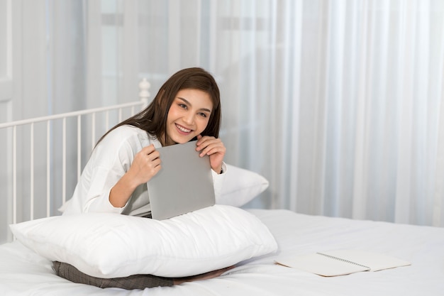 Glückliche lässige schöne asiatische Frau, die auf Laptop-Computer arbeitet, während auf dem Bett im Haus gesetzt wird.