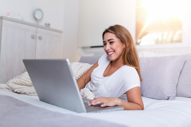 Glückliche lässige schöne Frau, die an einem Laptop arbeitet, der auf dem Bett im Haus liegt. Freiberuflerin, die zu Hause an ihrem Laptop arbeitet