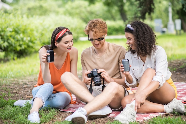 Glückliche Leute. Rothaariger Mann mit Sonnenbrille mit Kamera und zwei Freundinnen mit Getränken, die beim Picknick sitzen und das Foto in guter Stimmung betrachten