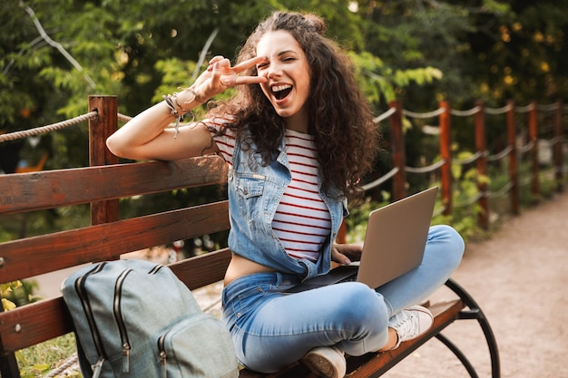 glückliche lustige Frau 20s mit lockigem Haar gestikuliert Friedenszeichen, während auf Bank im grünen Park sitzend, und studierend oder arbeitend auf silbernem Laptop