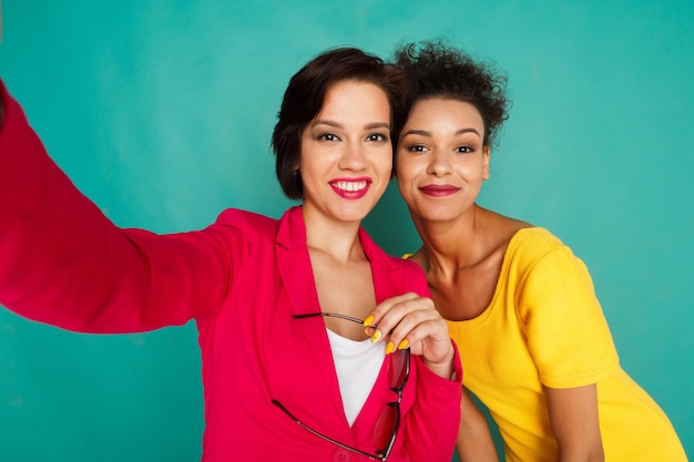 Glückliche mädchen, die selfie machen. lächelnde kaukasier- und mulattinmädchen in farbenfroher freizeitkleidung, die fotos auf blauem studiohintergrund machen, kopierraum. fotografie und technologie, multiethnisches freundschaftskonzept.