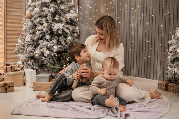 Glückliche Mama und Söhne sitzen auf dem Boden und unterhalten sich in einem Raum in der Nähe des Weihnachtsbaumes