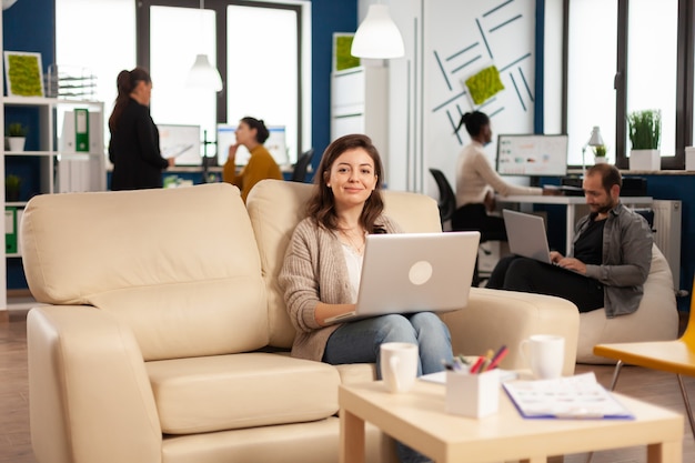 Glückliche Managerfrau, die auf dem Sofa vor der Kamera sitzt und den Laptop lächelt, während verschiedene Kollegen im Hintergrund arbeiten