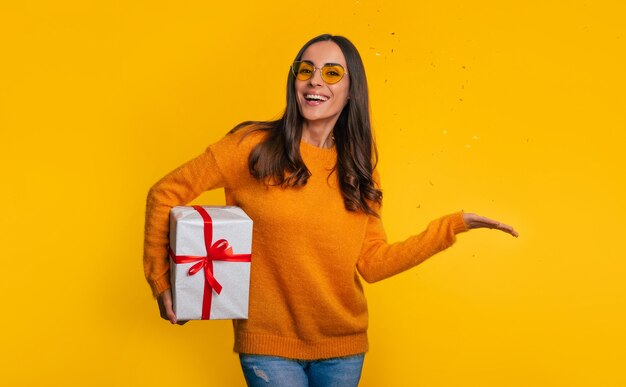 Glückliche moderne Frau in Gläsern mit Geschenkbox in der Hand schaut in die Kamera und lächelt