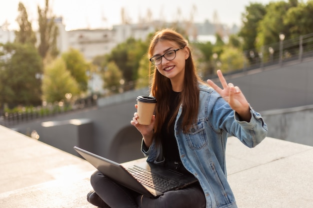 Glückliche moderne Studentin in einer Jeansjacke, die auf Treppe mit Laptop sitzt und eine Geste v. Fernunterricht zeigt. Modernes Jugendkonzept.