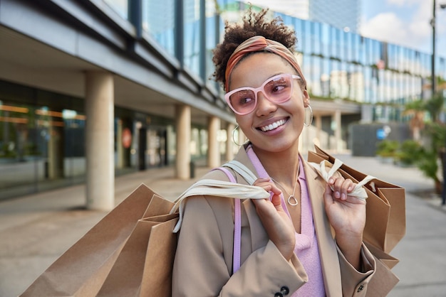 Glückliche modische Frau trägt ein trendiges Sonnenbrillenstirnband und eine Jacke, die froh ist, Einkäufe zu tätigen, trägt Einkaufstaschen