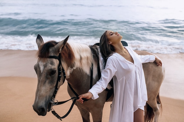 Glückliche modische junge Frau in einem weißen Kleid posiert mit einem Pferd am Strand.