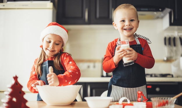 Glückliche Mühe und Schwester kochen etwas in der Küche, während sie sich auf die Weihnachtsferien vorbereiten, die Weihnachtsmannkleidung tragen