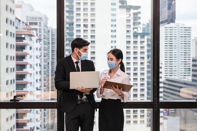 Glückliche multiethnische Kollegen, die Laptop und Zwischenablage halten und im Büro in der Innenstadt diskutieren. Tragen von Gesichtsmasken zum Schutz der Pandemie des Coronavirus, Covid-19