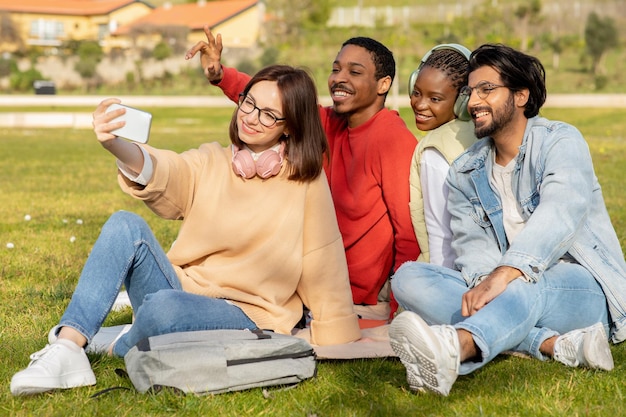 Glückliche multiethnische tausendjährige Studenten sitzen auf Gras und machen zusammen Selfies, um an der Universität zu bloggen