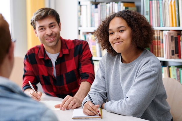 Glückliche multikulturelle Studentin in Freizeitkleidung, die einem der Teamkollegen während der Diskussion des Themas zuhört